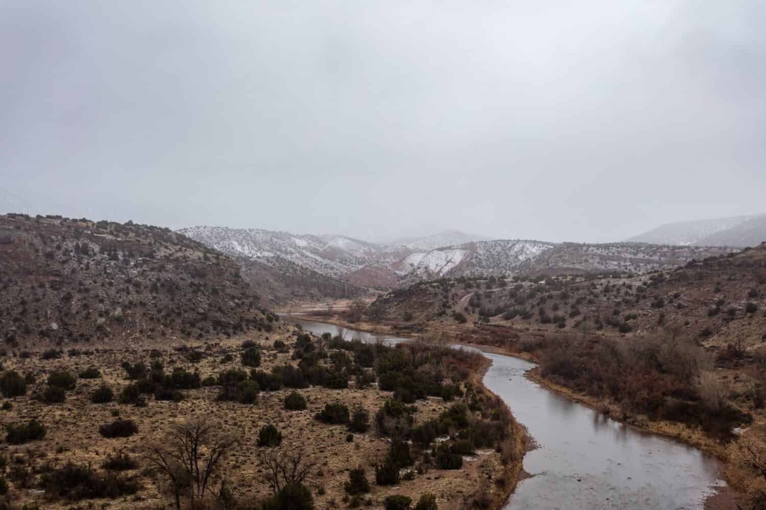 Rio Grande river cutting through large hills covered in bushes and snow on rainy overcast day in rural New Mexico