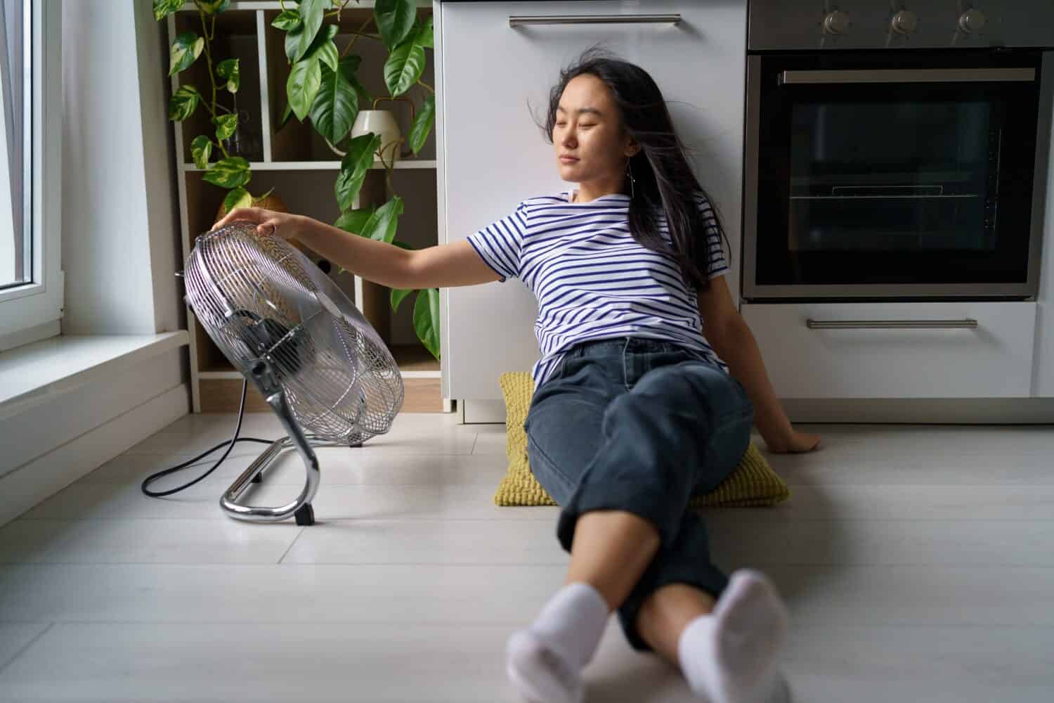 Overheating in homes. Tired overheated young Asian woman sitting on floor in kitchen near electric fan, cooling down at home during extreme summer heat, staying cool without air conditioning