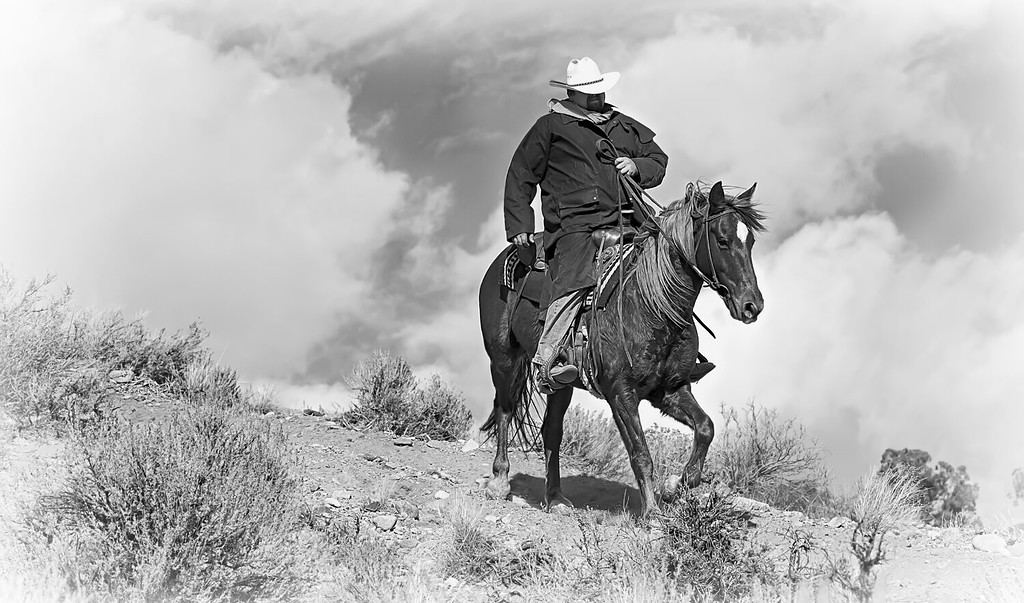 Pony Express Ridge Rider 1 in Black and White. A lone cowboy in a black coat rides his horse down a mountain ridge with dramatic skies behind him.