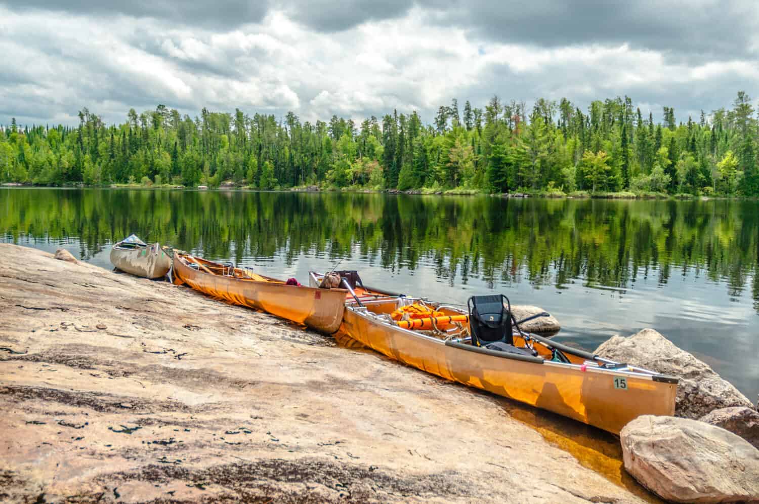 Three canoes line a rocky shore made of igneous rocks in the Boundary Waters Canoe area in the North Woods of Minnesota with beautiful green trees reflecting in the lake behind.
