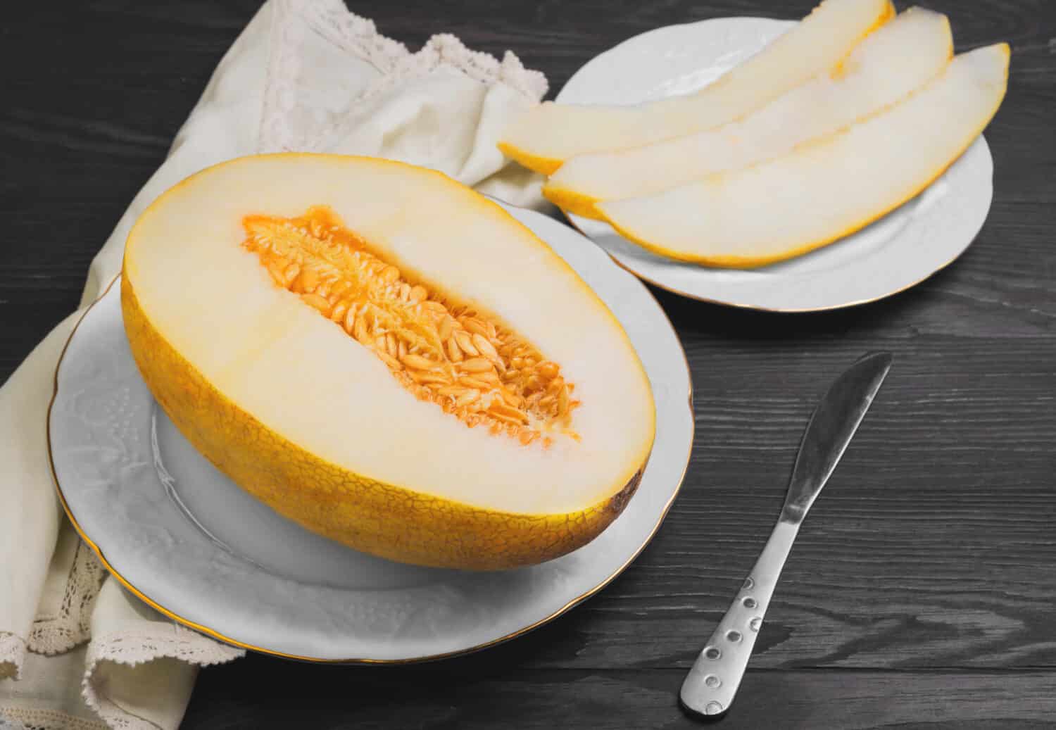 Fresh ripe yellow melon on white porcelain plate, three pieces of sliced melon, knife, cloth, dark brown wooden background