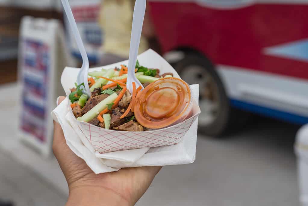 To go take away paper food tray of chicken teriyaki with chili sauce in deli containers, napkins, fork and spoon with blurry food truck background