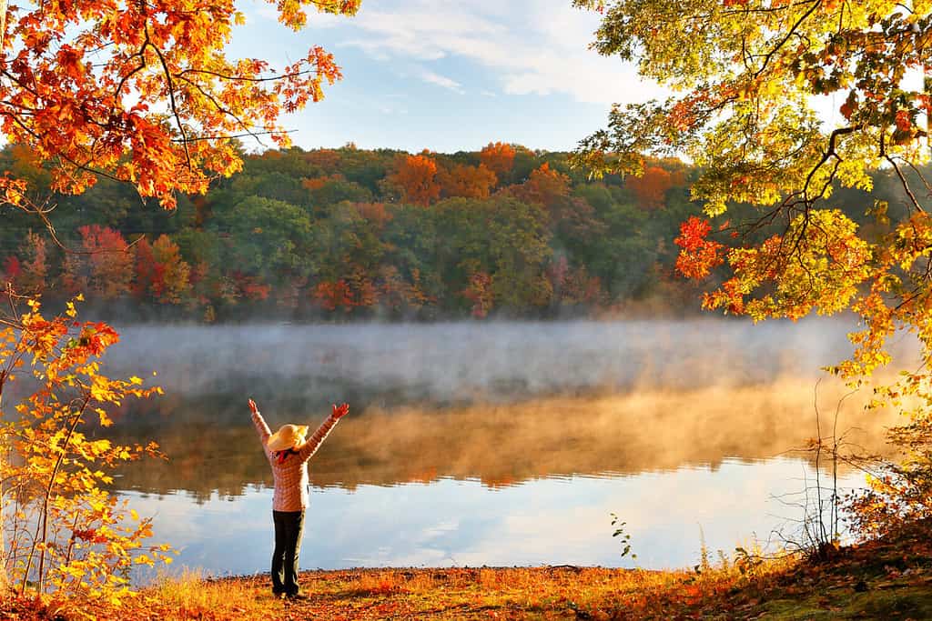 Beautiful Fall Foliage of New England at Sunrise, Boston Massachusetts. Photo shows a woman with a hat standing in front of foggy lake.