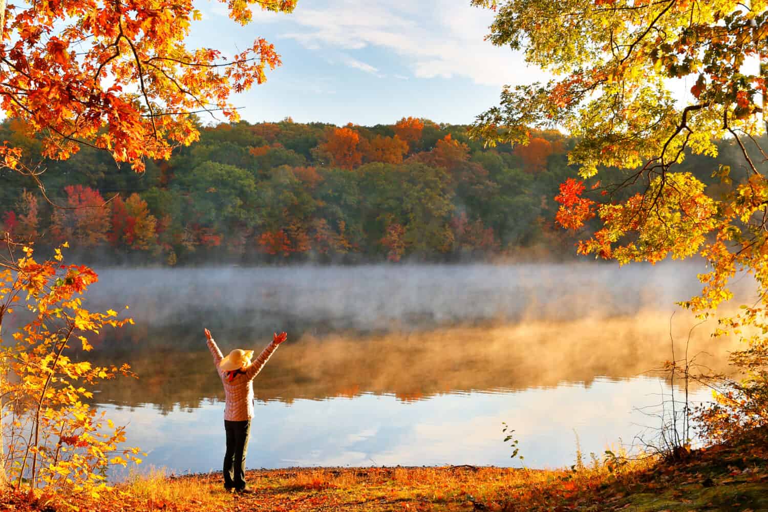 Beautiful Fall Foliage of New England at Sunrise, Boston Massachusetts. Photo shows a woman with a hat standing in front of foggy lake.