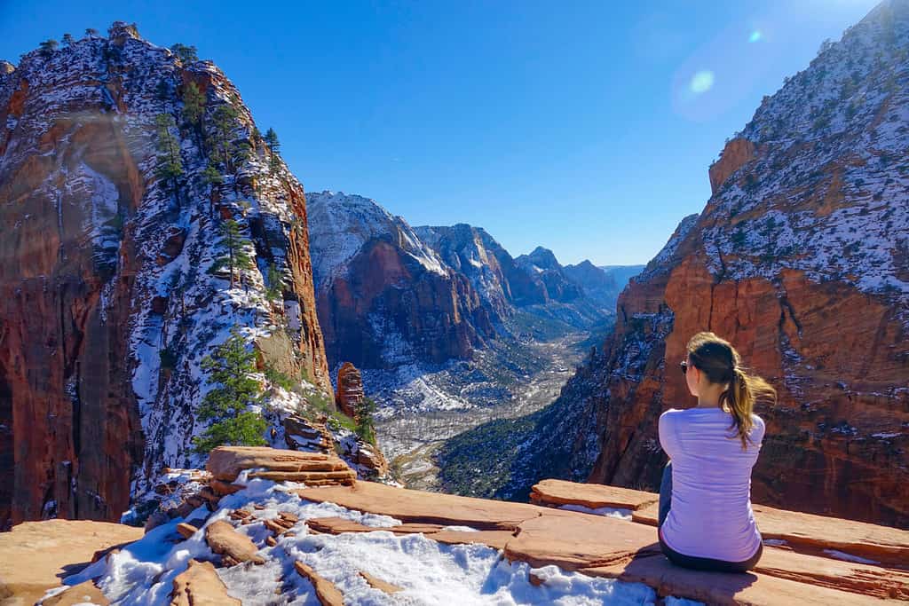 LENS FLARE: Traveler girl sits and observes the snowy canyon from Angel's Landing. Young woman sits down after a hike and looks at the beautiful red colored canyon in Zion National Park in Utah.