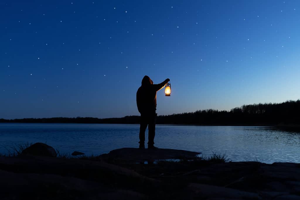 Man holding the old lamp outdoors near the lake. Hand holds a large lamp in the dark. Ancient lantern illuminates the way on a night. Light and hope concept.