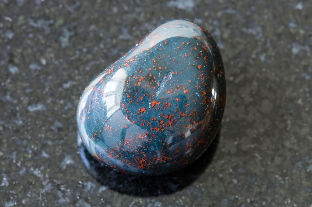 macro photography of sample of natural mineral from geological collection - tumbled Heliotrope (Bloodstone) gemstone on black granite background