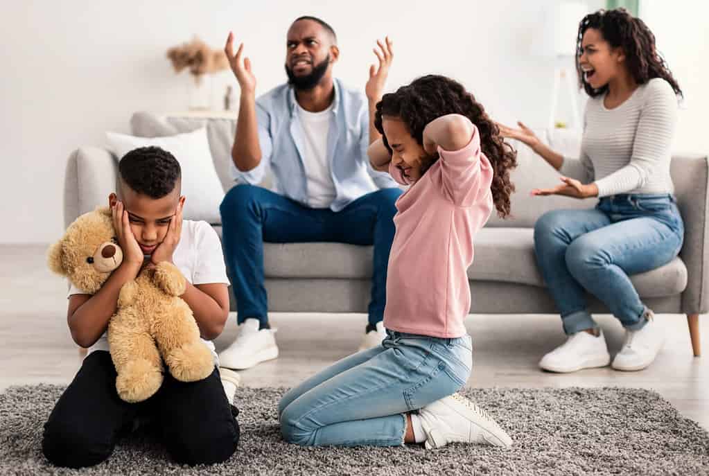 Family Conflicts. Sad little black children covering ears with hands while their parents arguing in the background, upset boy and girl don't want to hear quarrel, stressed kid sitting on the floor