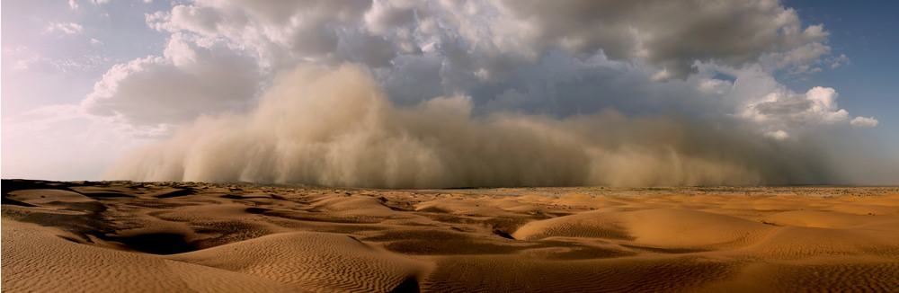 An image showing a severe sandstorm of high altitude with cumulonimbus rain clouds forming near towering mountains heading towards a sandy desert.