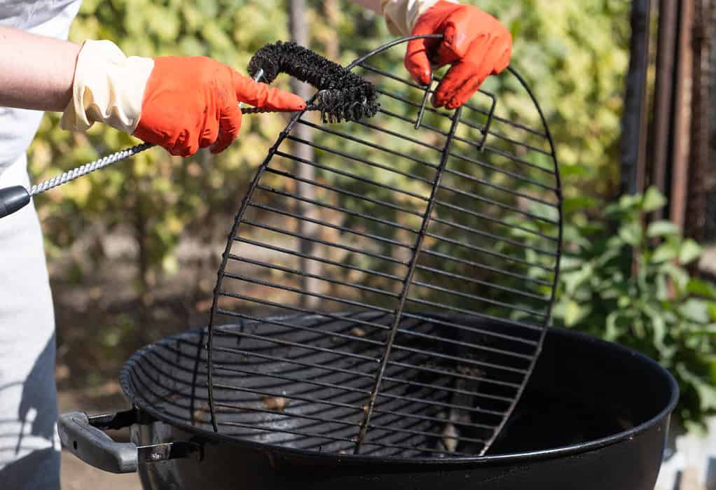 Male hand with red gloves cleans round grill with stiff brush.