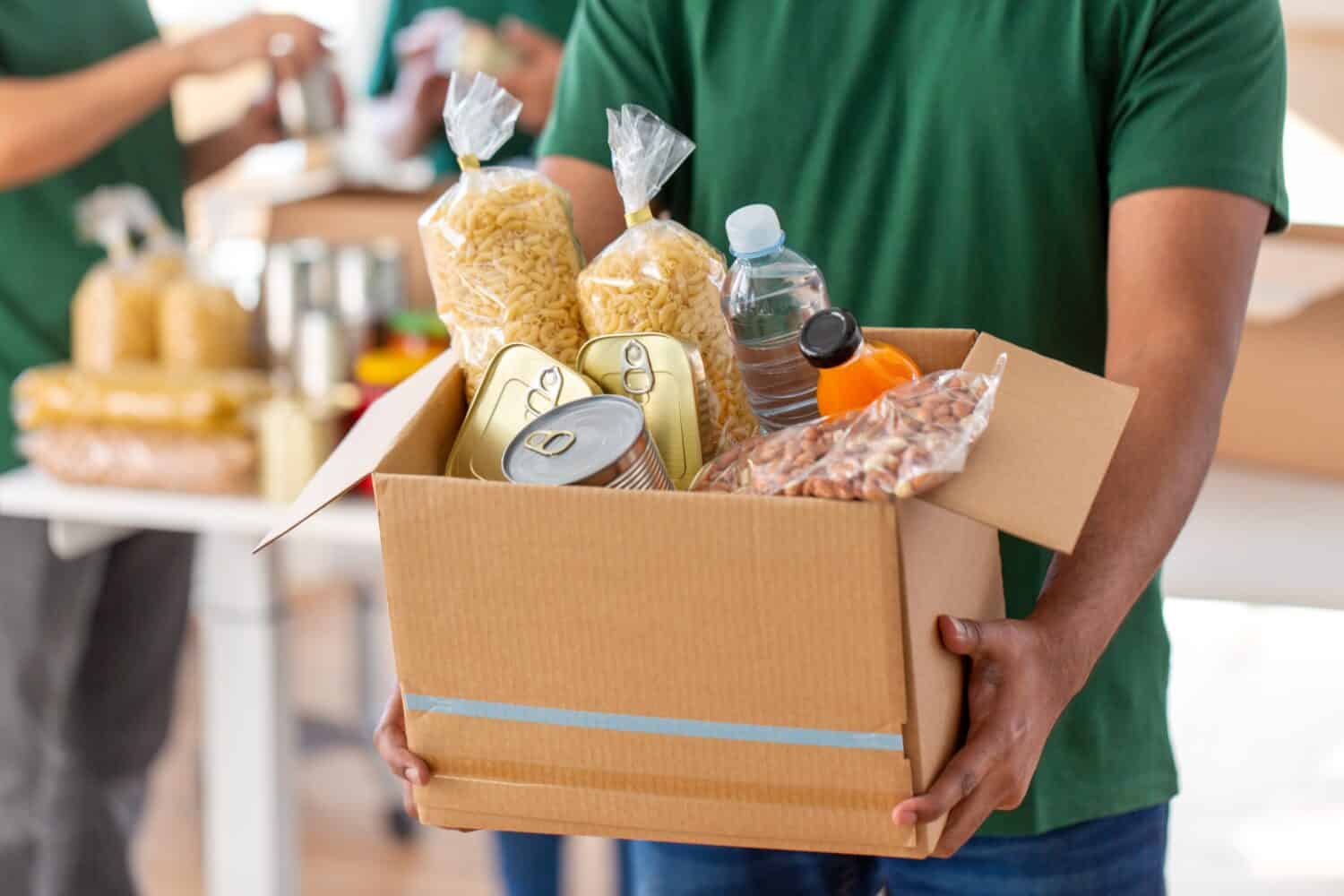 charity, donation and volunteering concept - close up of male volunteer's hands holding box with food over group of people at distribution or refugee assistance center