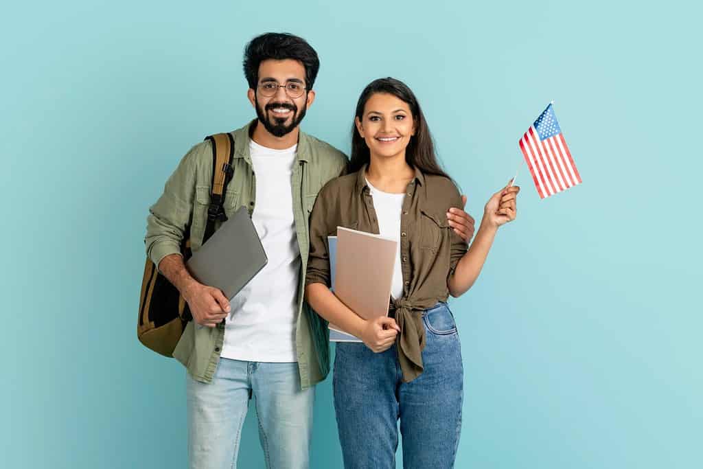 Cheerful happy beautiful multiethnic millennial couple arab man indian woman students with backpack, laptop and notepads showing flag of US, studying abroad, learning English, blue studio background