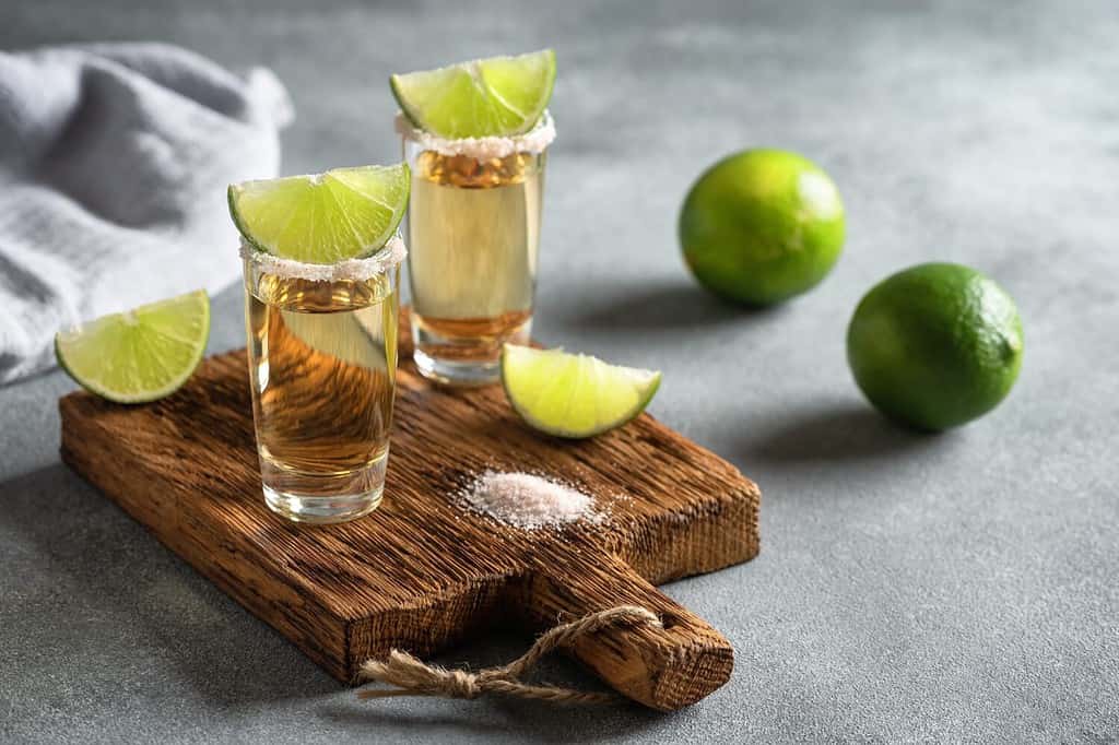 Mexican gold tequila in a shot glass with lime and pink salt on a wooden board, gray concrete background. Side view, selective focus.