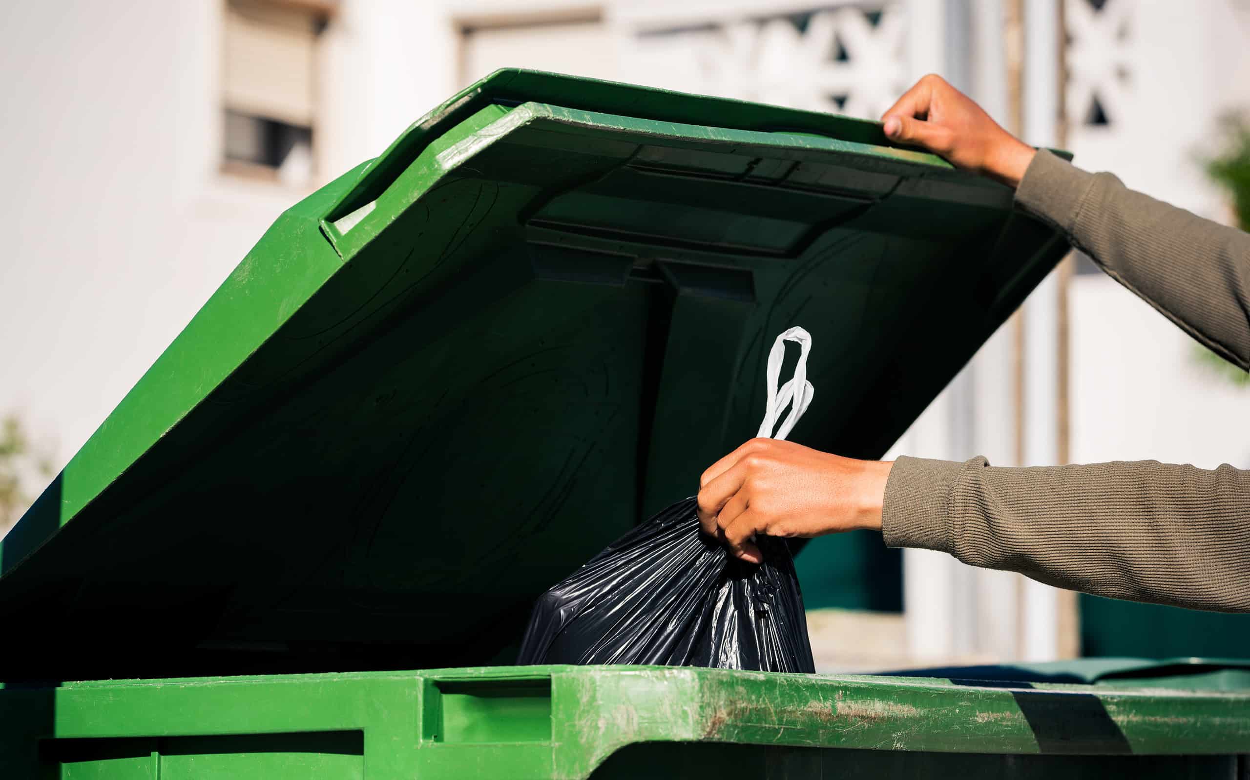 Man throwing out black eco-friendly recyclable trash bag in to big plastic green garbage container. Take out the trash