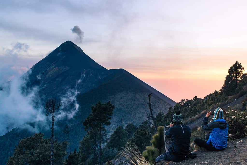 Two young hiker men contemplating the view from the Acatenango Volcano camp area where the Fuego Volcano partially covered in fog can be seen on a picturesque afternoon with dense forest