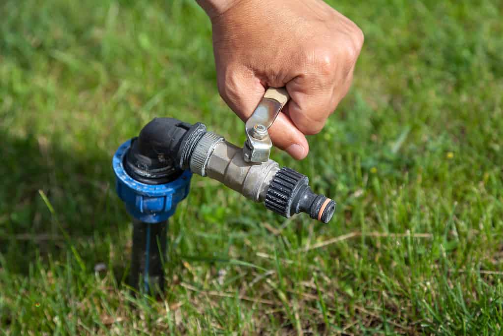 Hand opens tap of water irrigation on green lawn