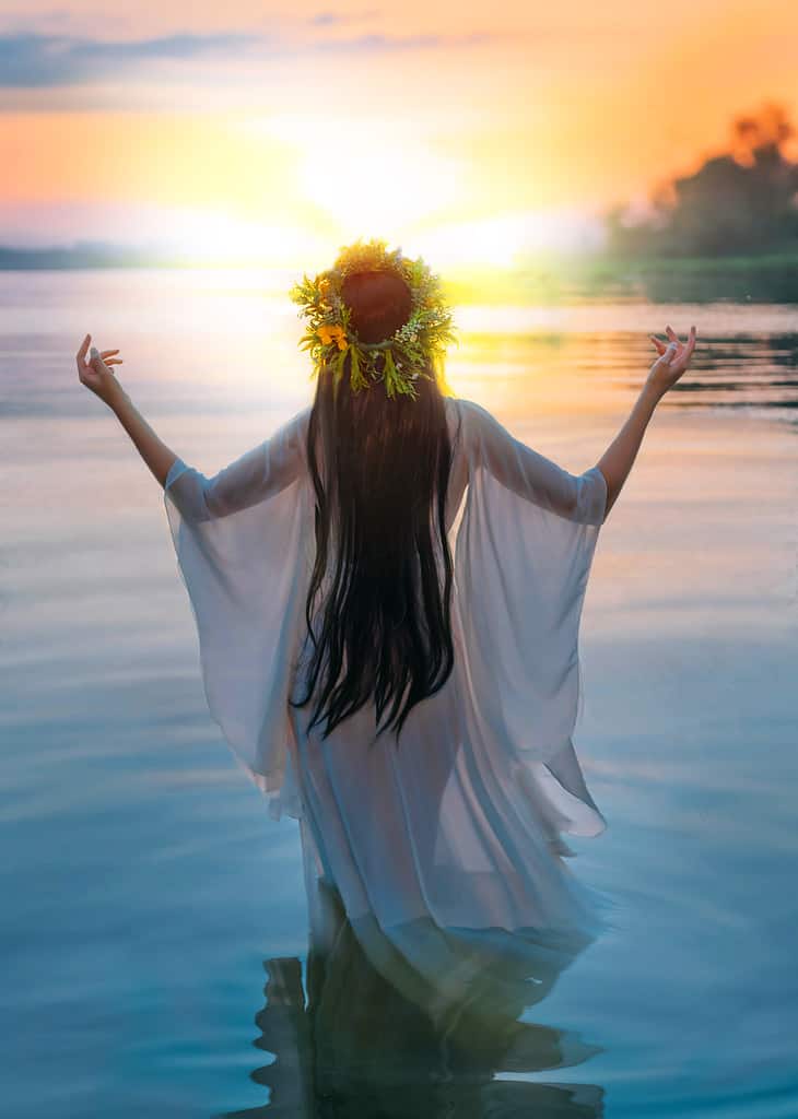 Photo with noise. Fantasy woman standing in water hands raised to sky, praying to sun. Slavic girl in herbal wreath, long hair white dress, pagan divination ritual. Nature blue river sunset. Back view