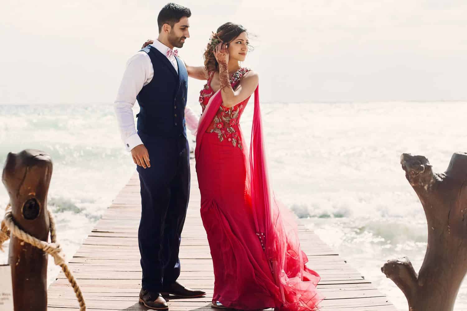 Wind blows bride's pink sari while she stands with Hindu groom on a wooden quay among foaming waves