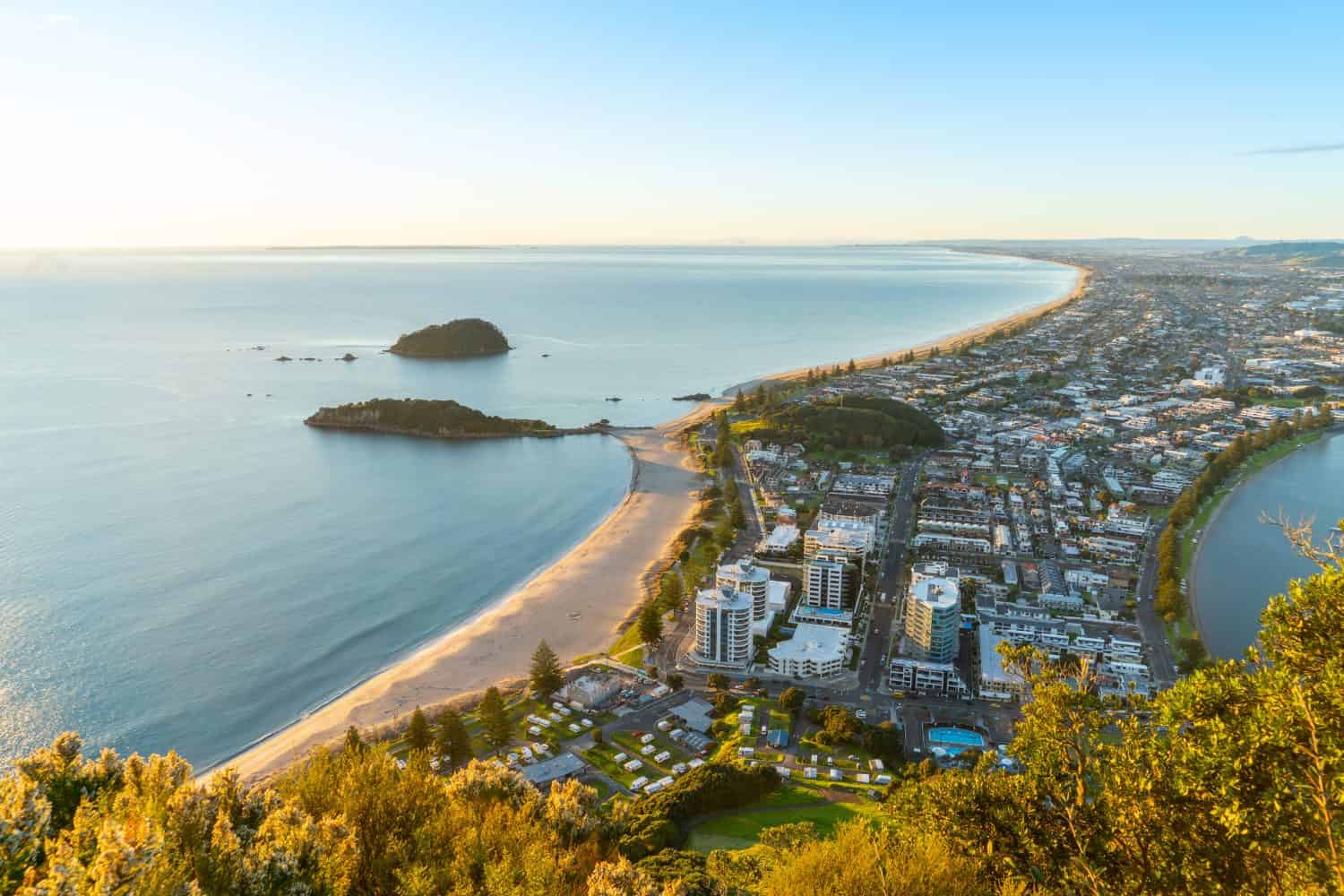 Mount Maunganui stretches out below as sun rises on horizon and falls across ocean beach and buildings below
