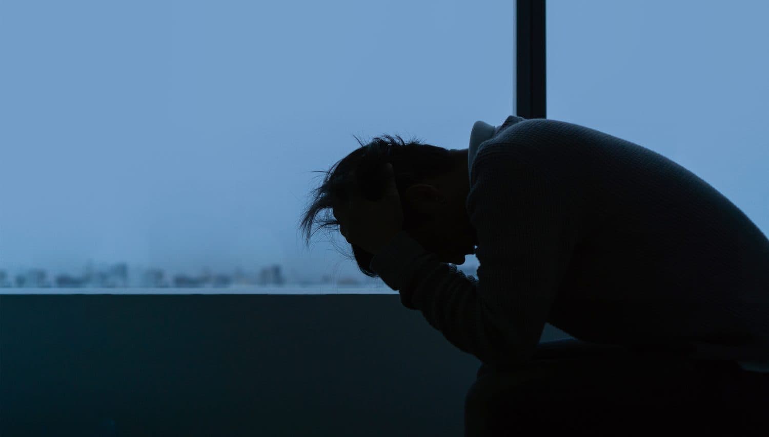 PTSD Mental health concept, Young depressed asian man sitting alone near window in evening time with low light environment, Copy space, selective focus.