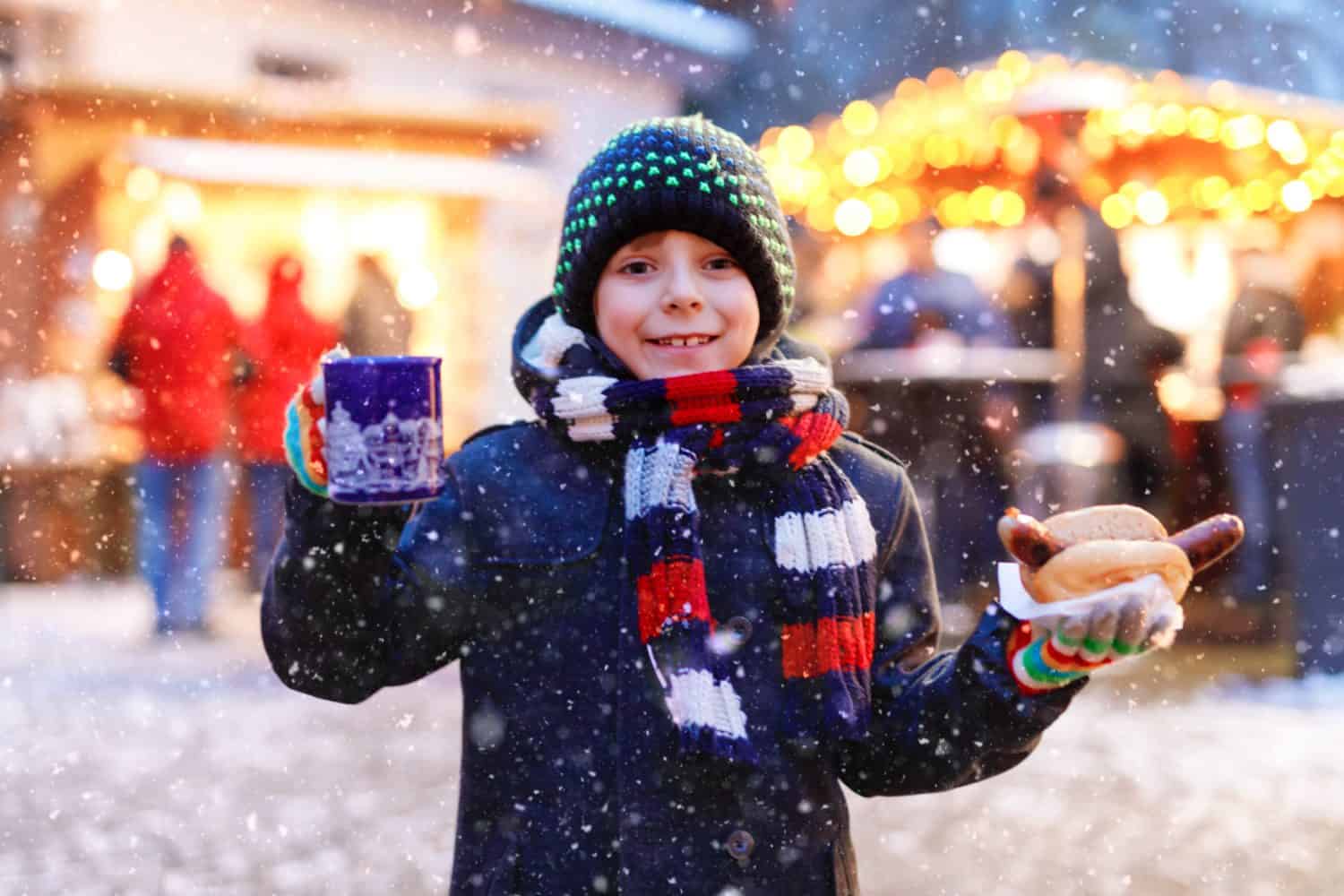 Little cute kid boy eating German sausage and drinking hot children punch on Christmas market. Happy child on traditional family market in Germany. Laughing boy in colorful winter clothes