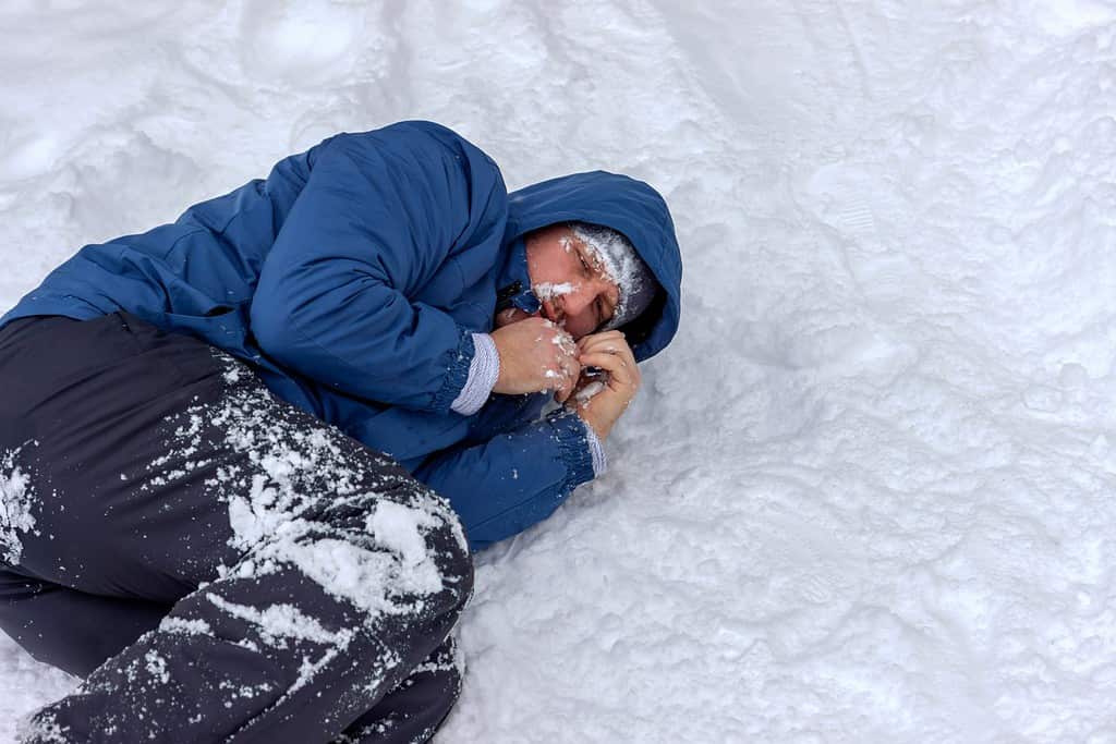 Frozen man in a blue jacket and hat lying down covered snow and frost, trying to stay warm on a very cold winter day, snow falls around him. Sick mountaineer with hypothermia on snow during the day.