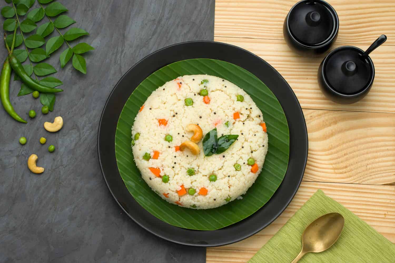 Upma made of samolina or rava upma, most famous south indian breakfast item which is arranged in a  black plate  and garnished with fried cashew nut and curry leaves with grey colour background.