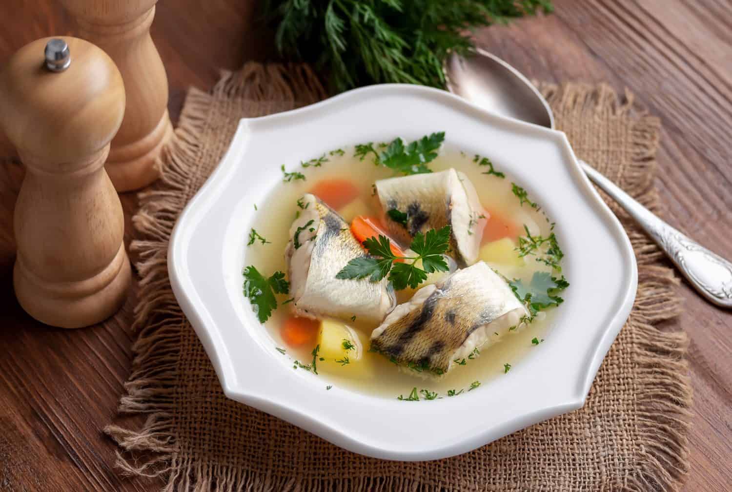 Russian Fish Soup Ukha. White fish in a clear broth with diced root vegetable served in white plate on wooden table. Selective focus.