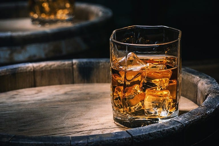 Bottle and glass of whiskey with ice on a wooden background. Glass of Scotch whiskey and ice sits on top of a rustic whiskey barrel. Whiskey with ice.