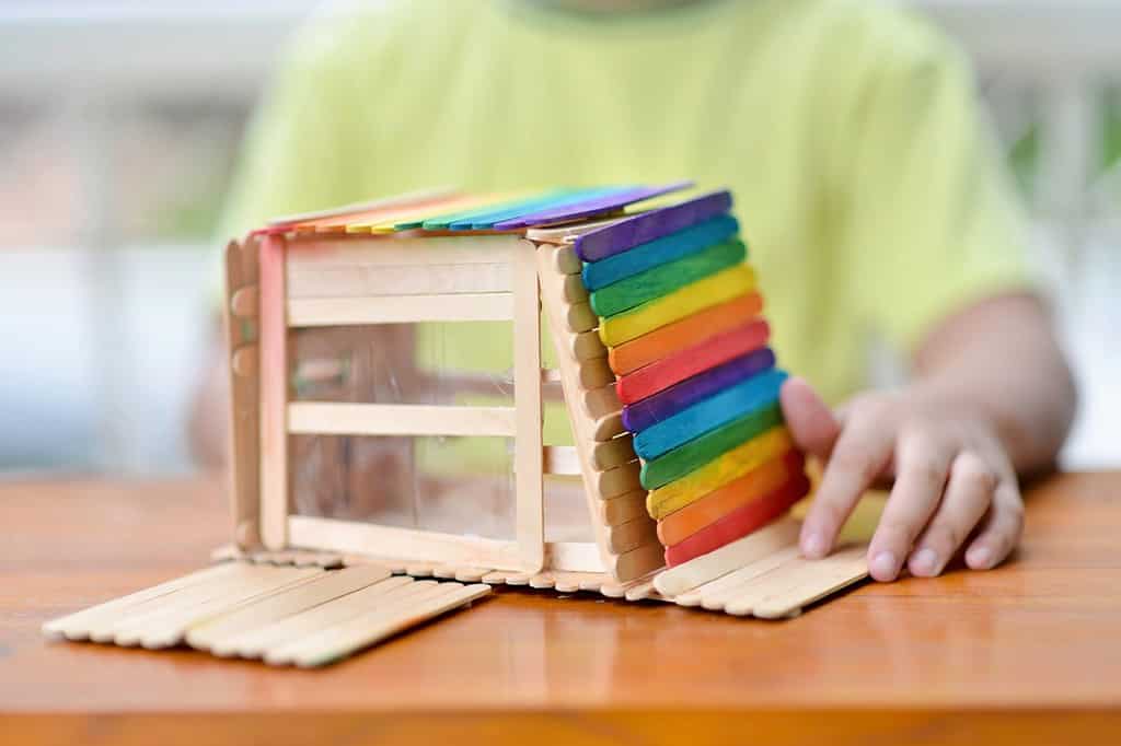 Asian child with popsicle sticks house in the outdoor.Young male with creative wooden innovation.Kid doing colorful model design craft.Homeschooling boy handmade activity project.