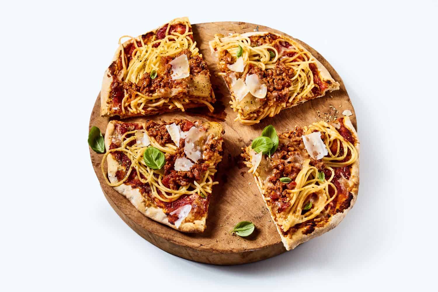 From above of delicious homemade pizza with minced meat and spaghetti garnished with basil and Parmesan cheese on white table
