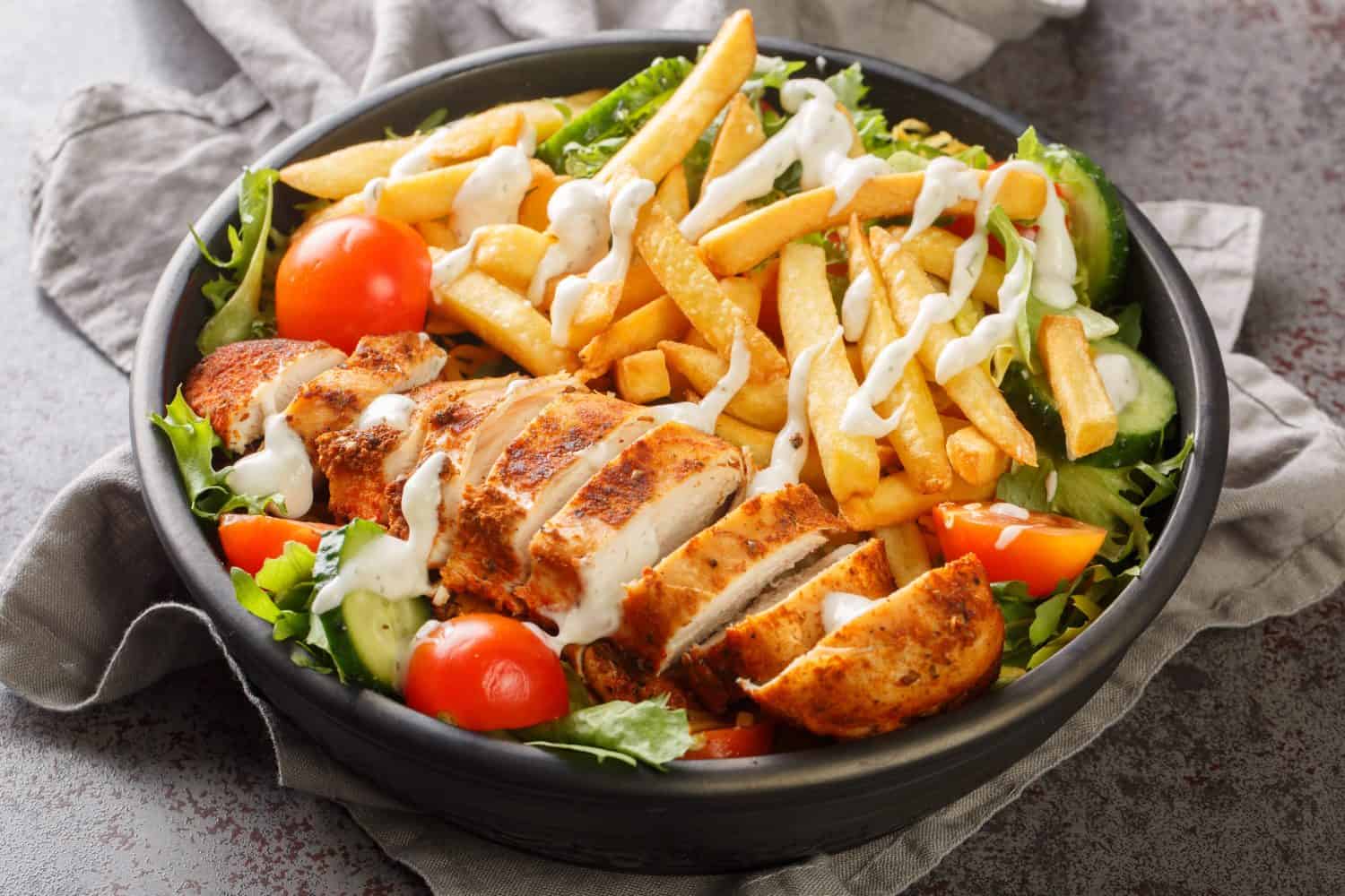 Pittsburgh steak salad consists of a bed of lettuce that's topped with tomatoes, cucumbers, a piece of grilled chicken, and crispy french fries closeup on the bowl on the table. Horizontal