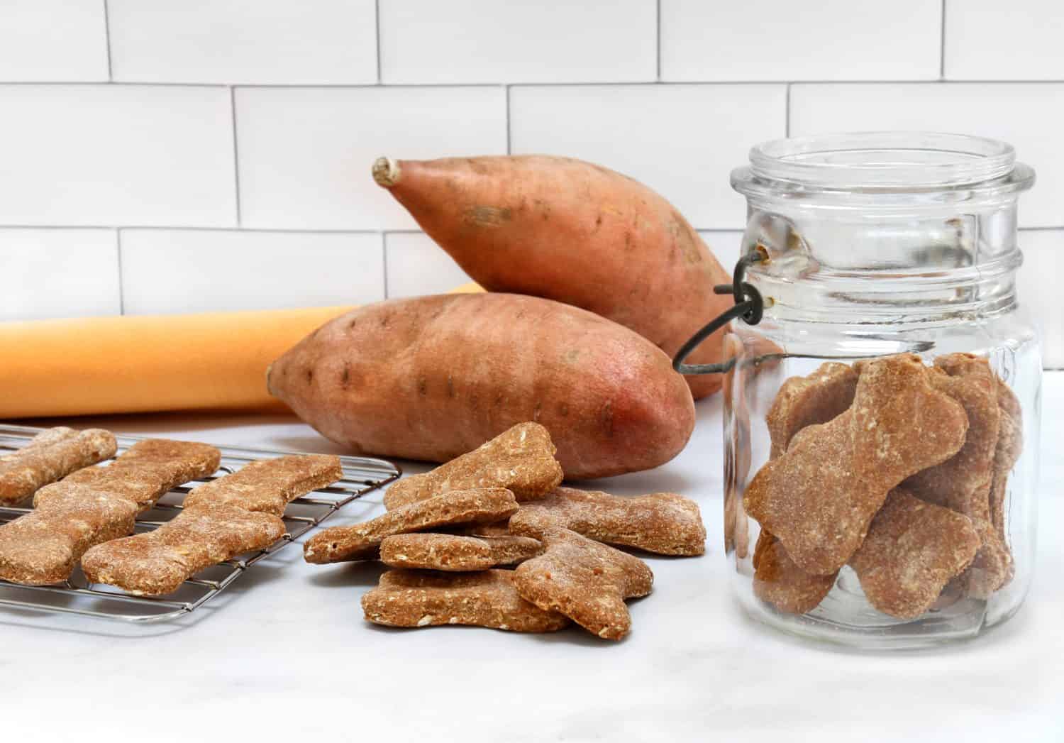 Dog cookies made from sweet potatoes on a rack, in a jar and stacked. Sweet potatoes in background.