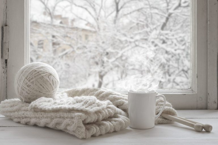 Cozy winter still life: mug of hot tea and warm woolen knitting on vintage windowsill against snow landscape from outside.