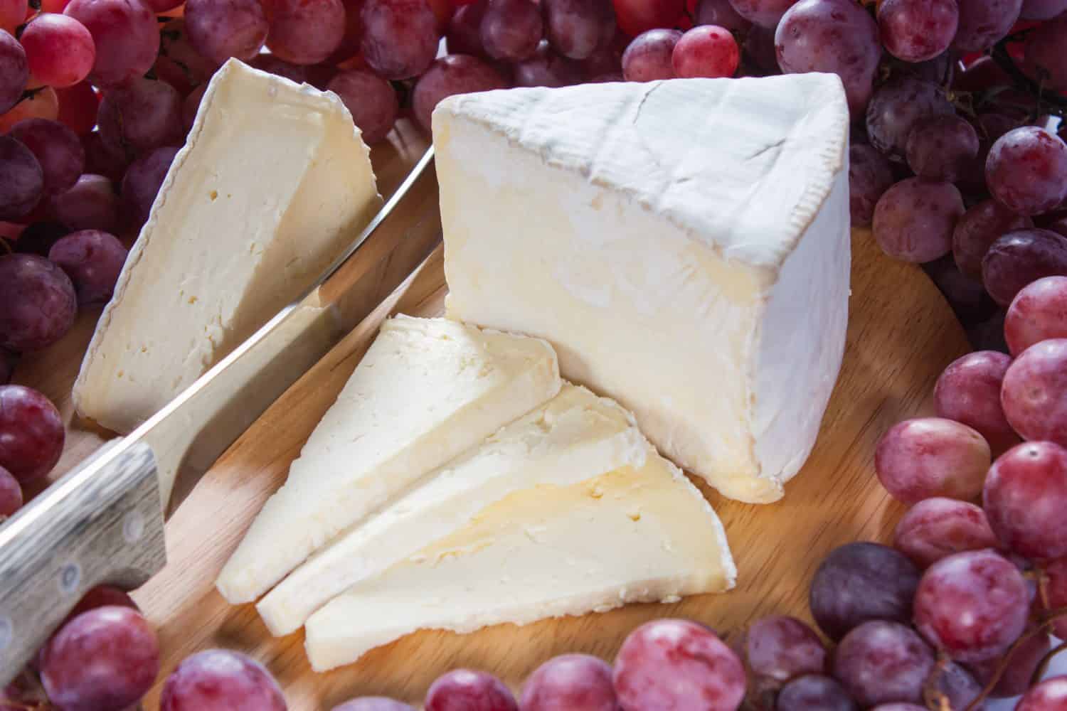 French cheese with white noble mold St. Andre on a cutting board with a knife surrounded by red grapes, horizontal