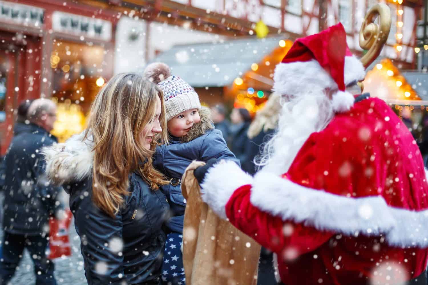 Cute toddler girl with mother on Christmas market. Funny happy kid taking gift from bag of Santa Claus. holidays, christmas, childhood and people concept. Happy family during snowfall on winter day.