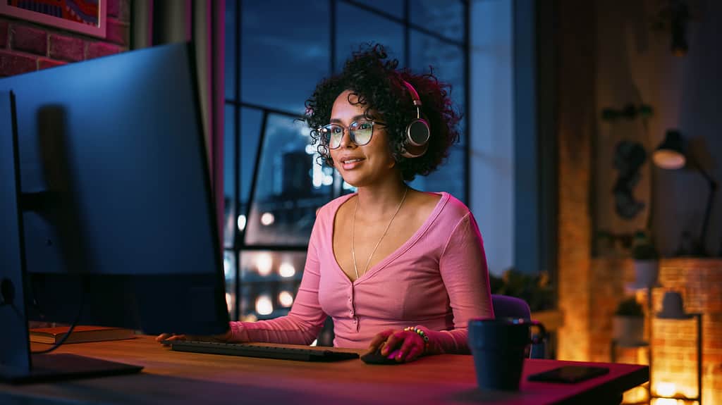 Young Beautiful Black Woman in Headphones Using Computer in Stylish Loft Apartment in the Evening. Creative Female Smiling, Browsing Videos on Social Media. Urban City View from Big Window.