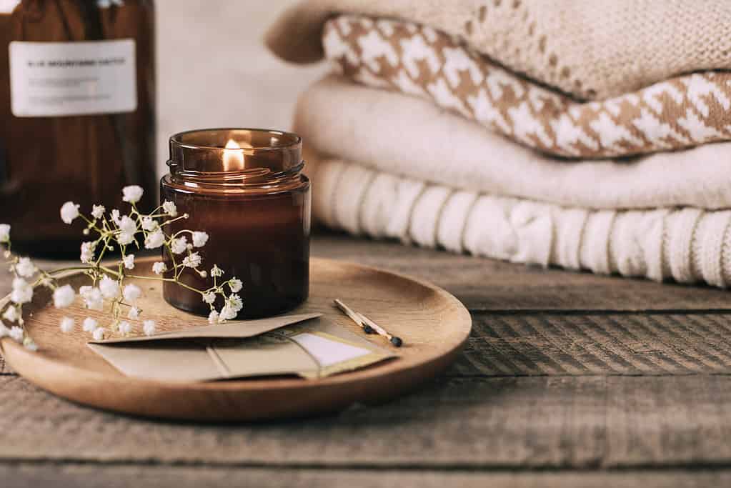 Burning candle in small amber glass jar, flowers of gypsophila and stack knitted seasin sweaters. Cozy lifestyle, hygge concept