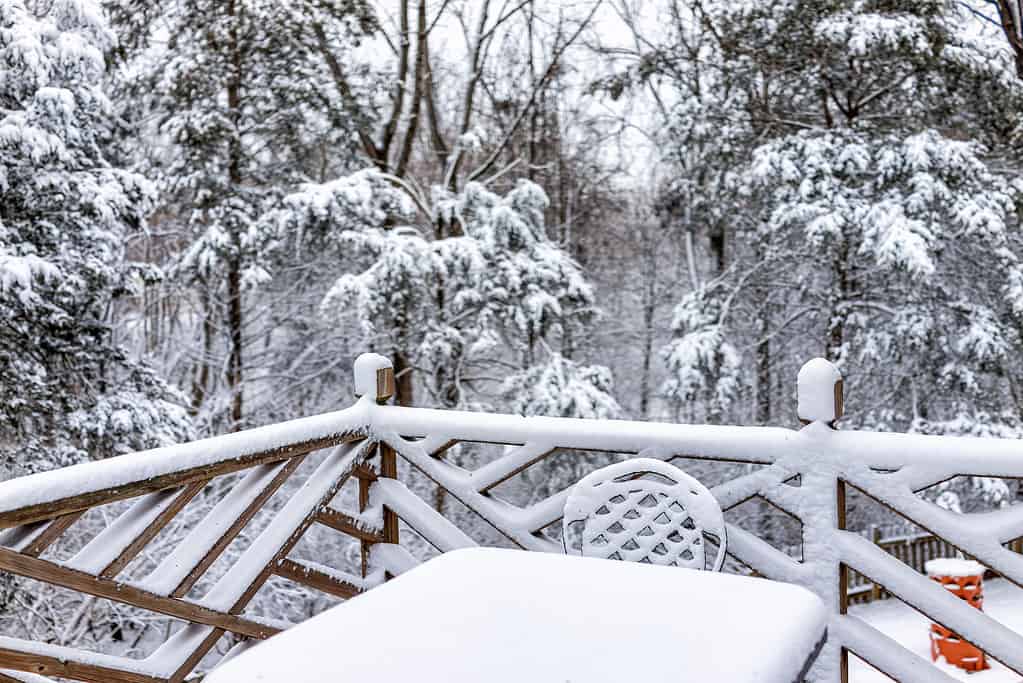 White snow covered metal outdoor table chair closeup on home house wooden deck with railing fence in winter with forest trees in northern Virginia
