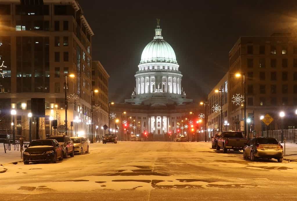 Madison, the capitol of Wisconsin downtown street view with parked cars and Wisconsin state capitol building glowing in the blizzard night. Wisconsin state, Midwest USA. Beautiful snowy winter night.
