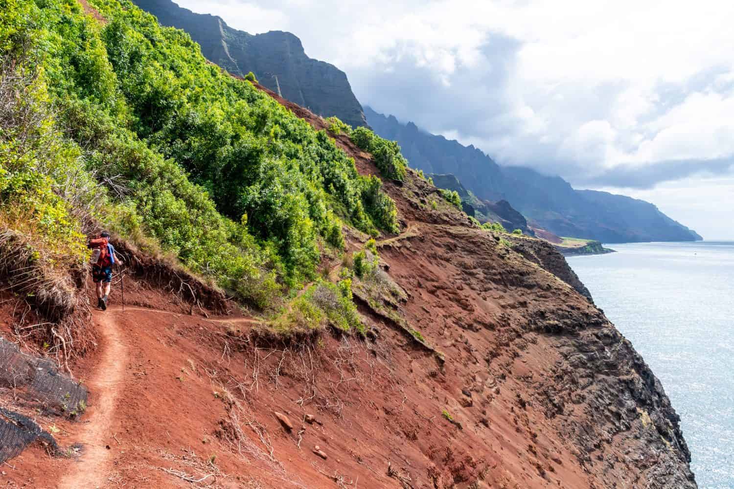 Hiker on a treacherous coastal trail with valleys and cliffs on one side and the ocean on the other. Shot on the Kalalau Trail in the Na Pali Coast State Park on the island of Kauai, Hawaii USA.