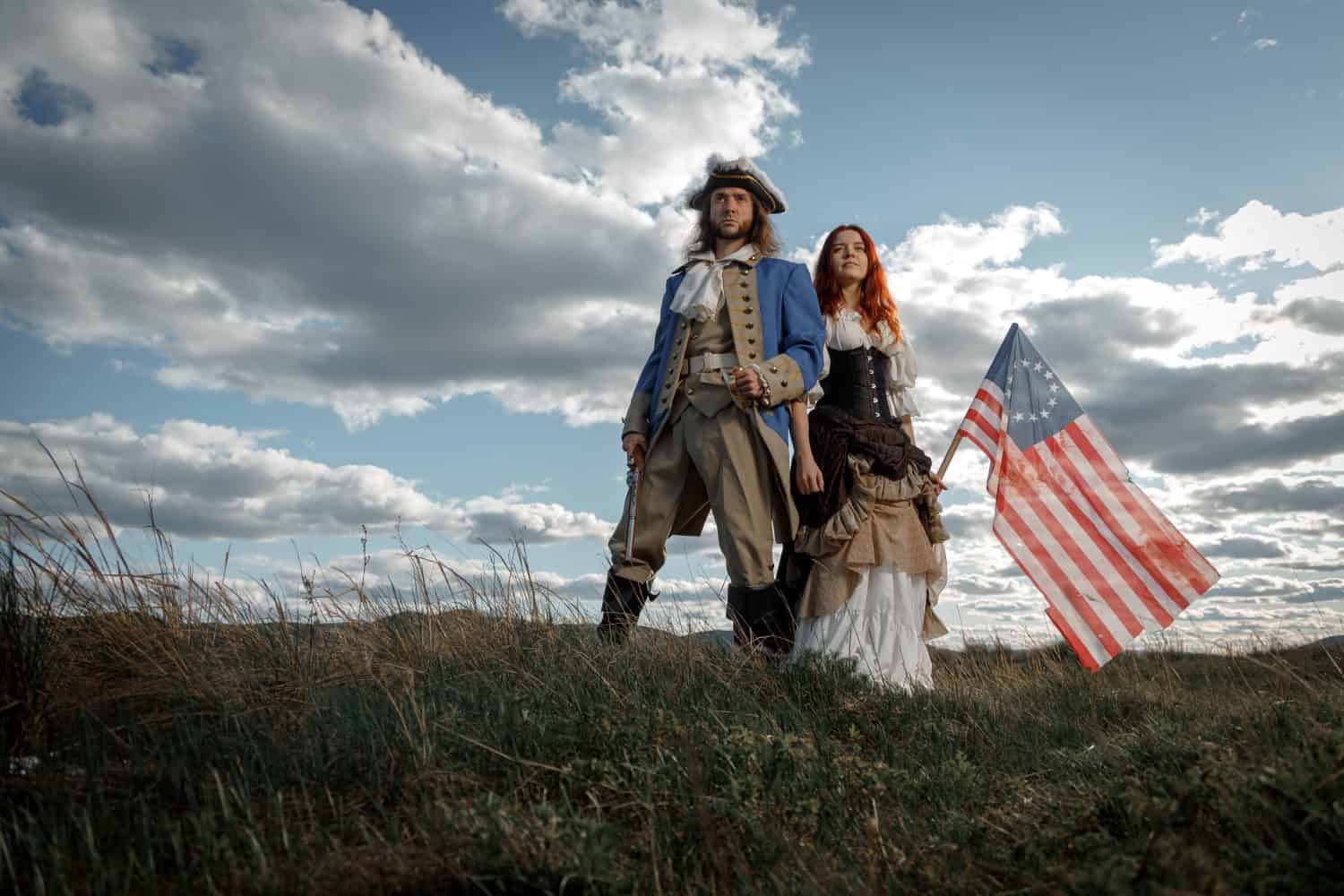 Man in form of officer of War of Independence and girl in historical dress of 18th century. July 4 is US Independence Day. Couple of patriots freedom fighters in outdoor on background cloudy sky. American Revolution. Revolutionaries.