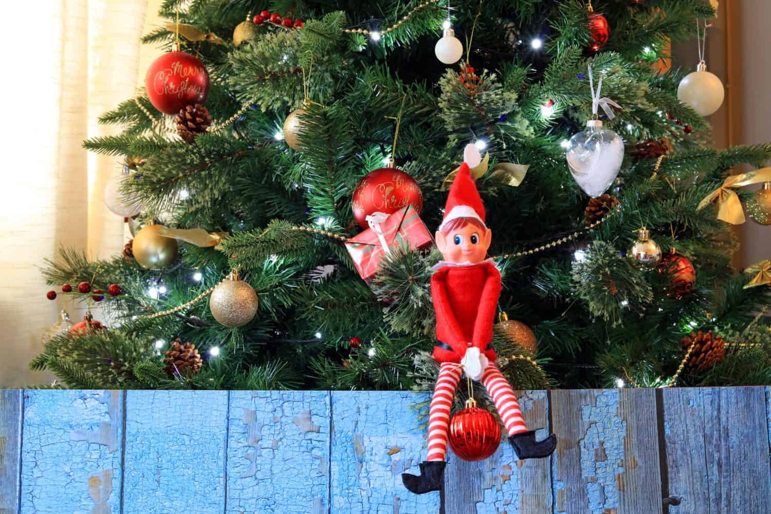 Mischievous Elf On The Shelf Sat On A Christmas Tree Branch Holding A Bauble.