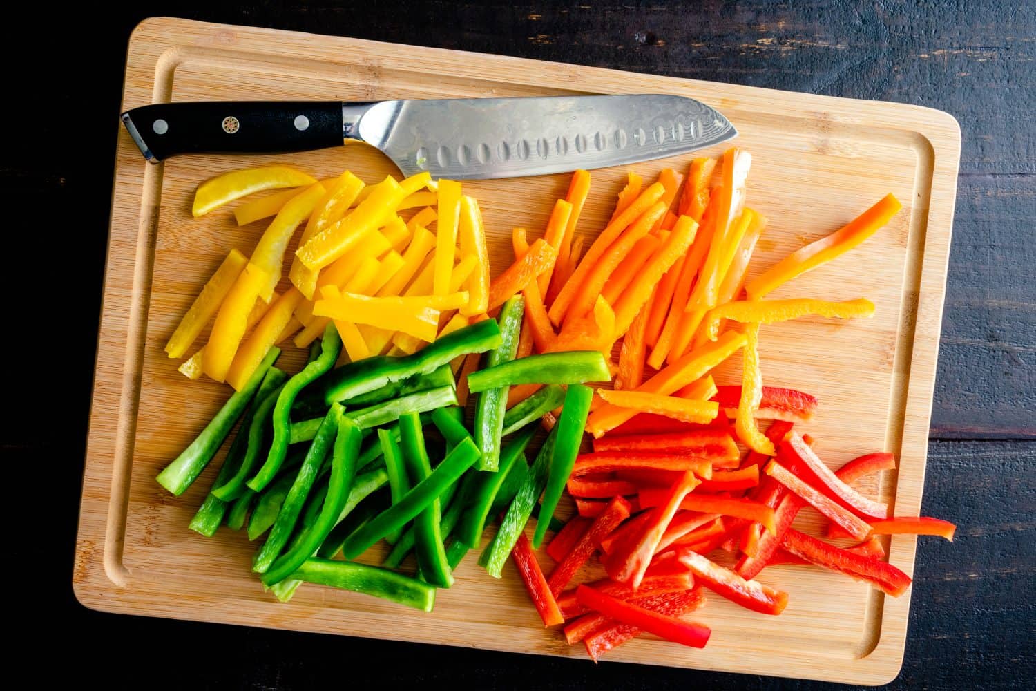 Red, Orange, Yellow, and Green Bell Peppers Cut into Thin Strips: Bell peppers cut in batonnets on a bamboo cutting board with a chef's knife