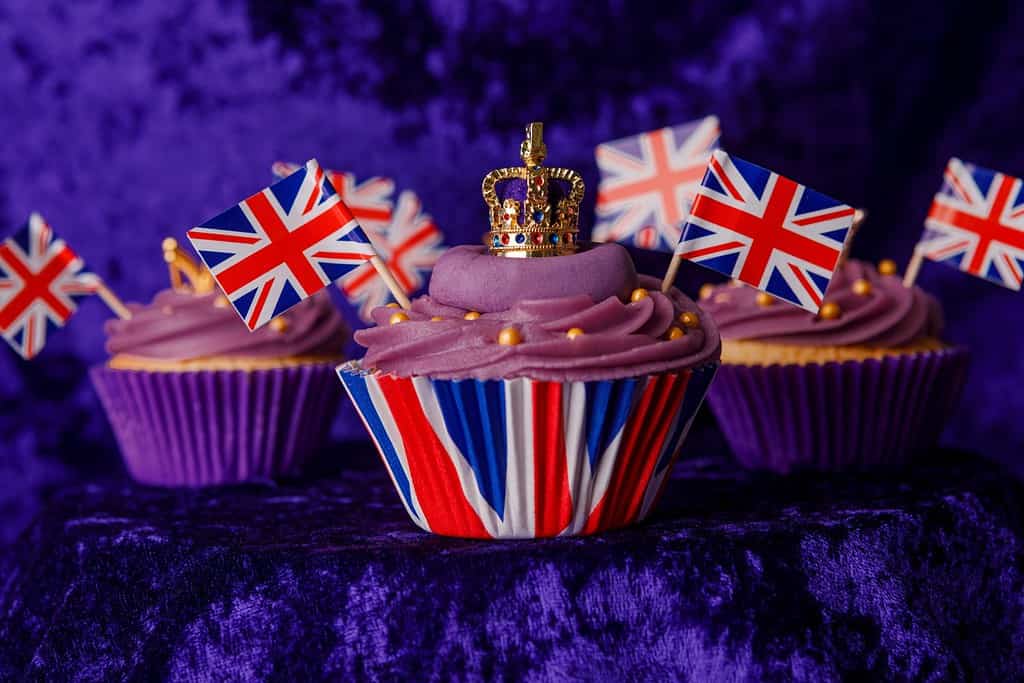 Royal Coronation Cupcakes to celebrate the coronation of King Charles III. Cupcakes decorated with the crown, purple velvet backdrop, union jacks flags, luxury cupcakes on a pedestal.