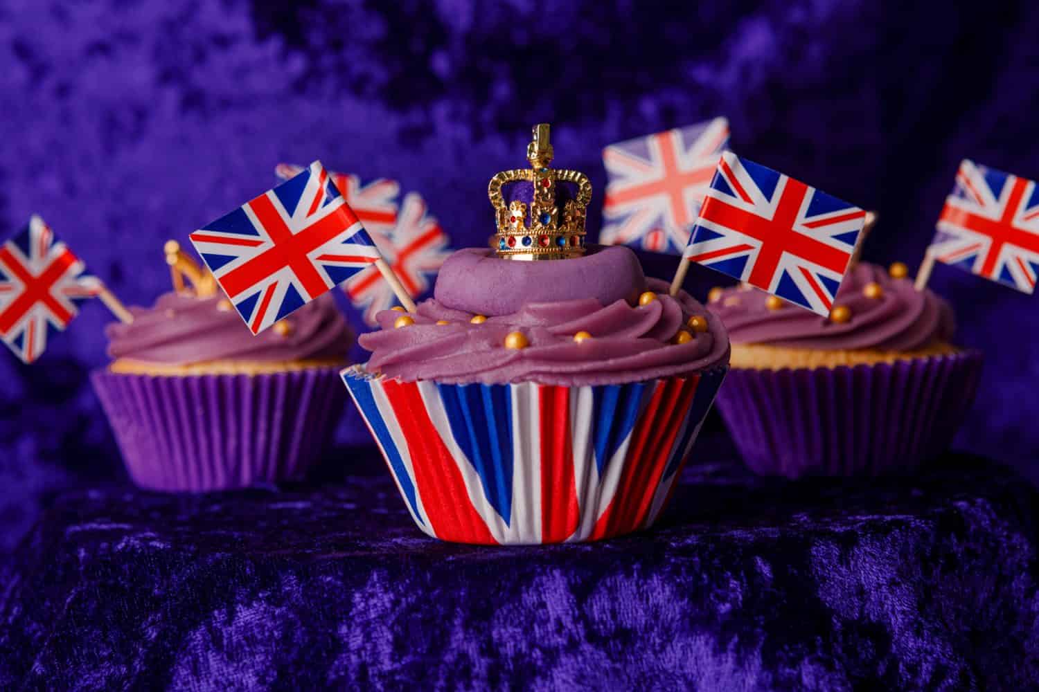 Royal Coronation Cupcakes to celebrate the coronation of King Charles III. Cupcakes decorated with the crown, purple velvet backdrop, union jacks flags, luxury cupcakes on a pedestal.