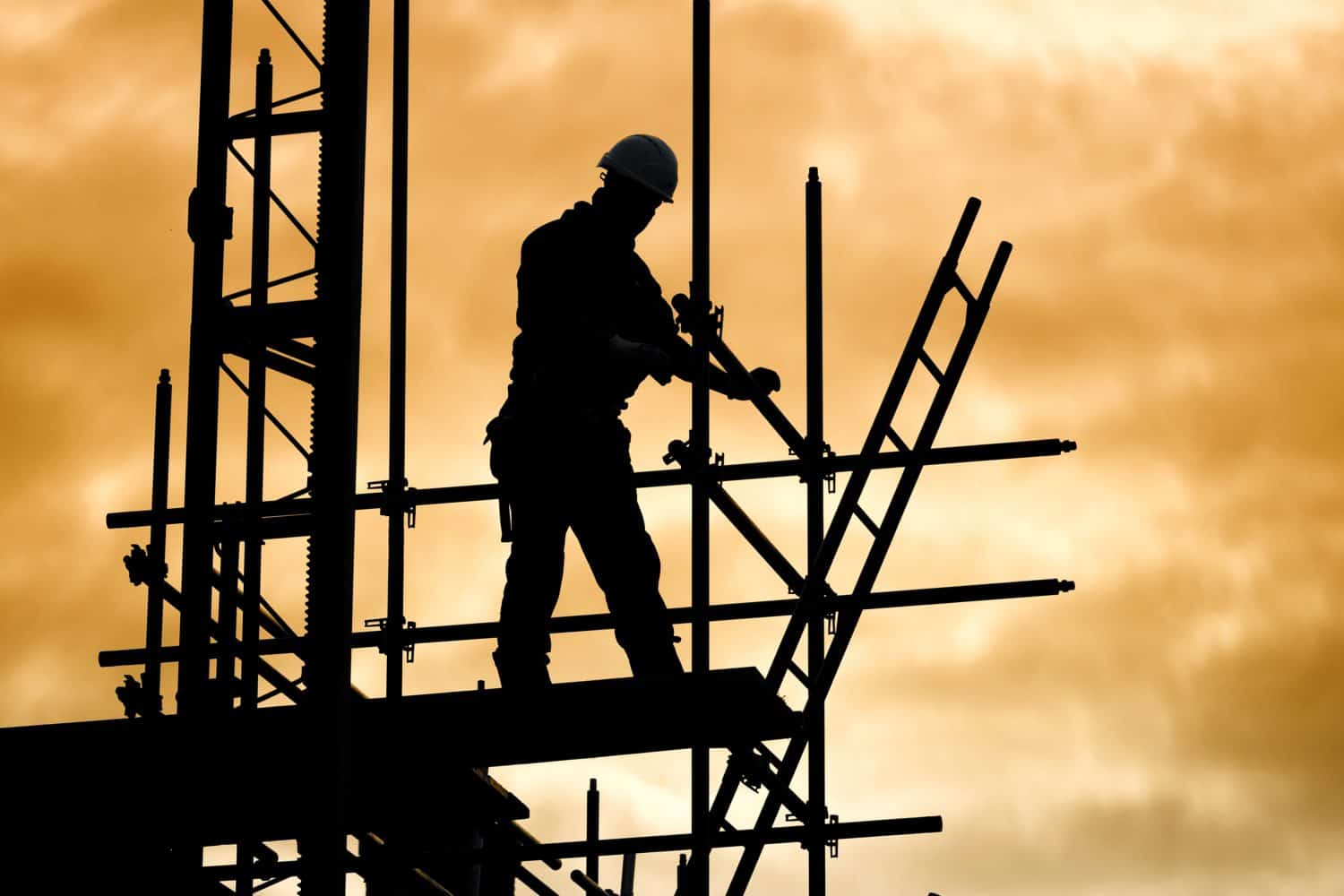 silhouette of construction worker against sky on scaffolding with ladder on building site at sunset