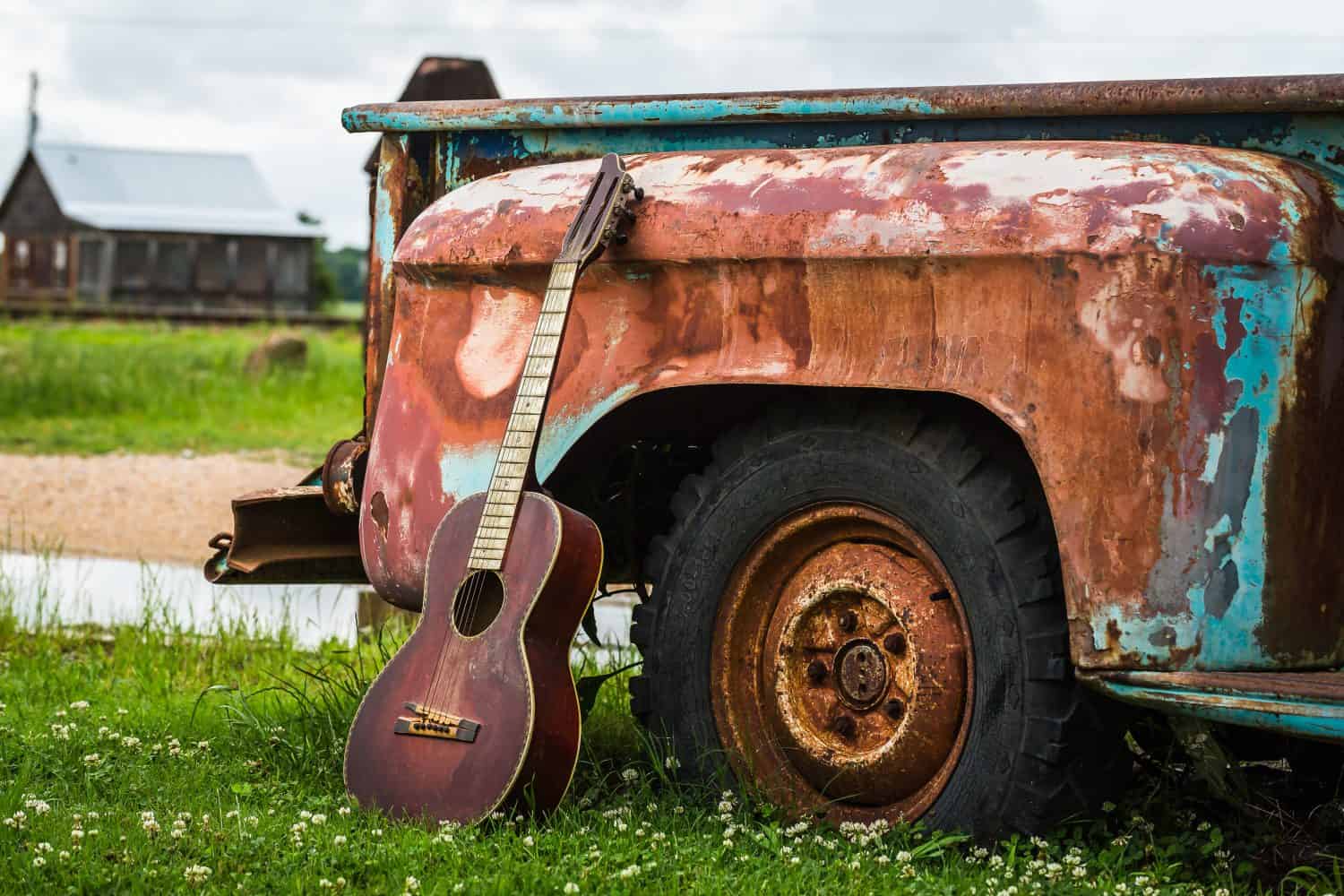 Color image of an old acoustic guitar leaning up against an old rusted truck parked in the grass with an old wood shack in the background in Clarksdale Mississippi