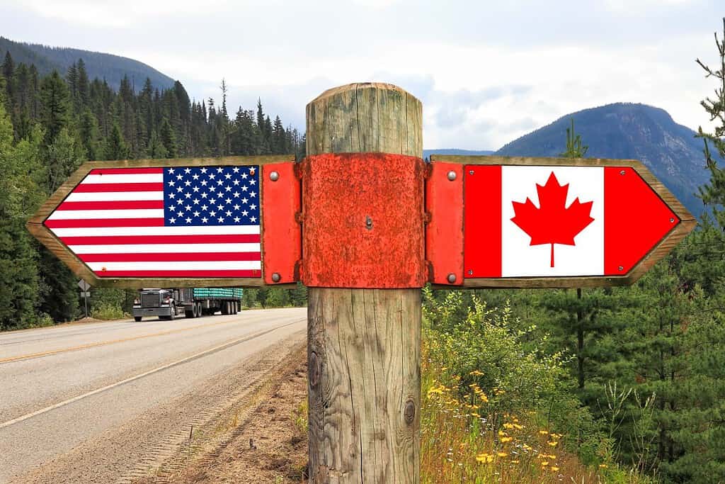Wooden signpost with two arrows - American and Canadian national flags on the highway road with nature landscape in the background. Canada and America moving direction sign