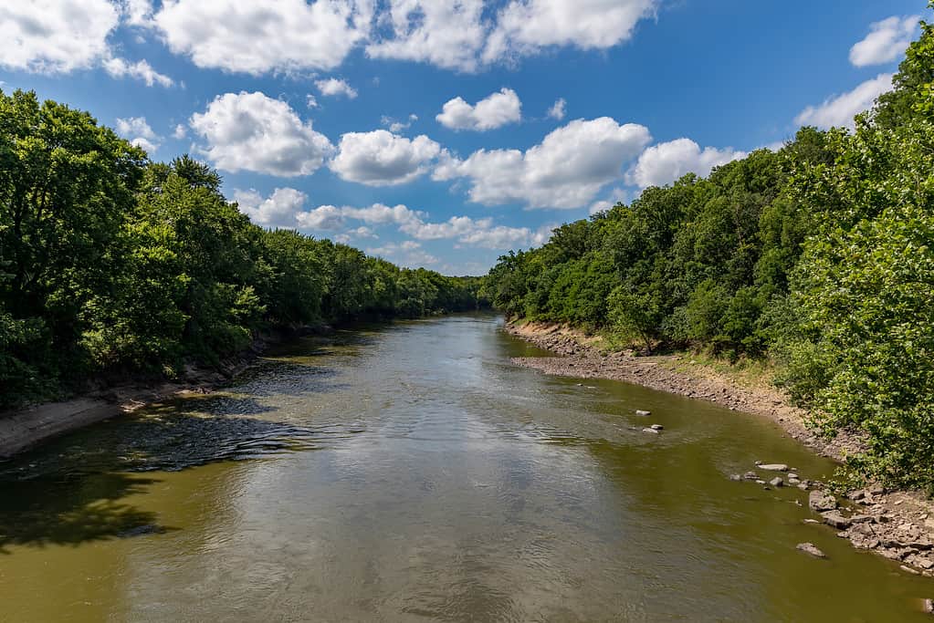 Water flowing along rocky riverbank of Sangamon River surrounded by trees on a sunny day with cloudy sky